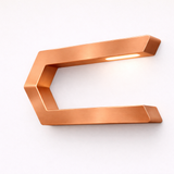 Copper finish C-shaped LED lamp by WOOM, with a brushed metal texture and glow from the top edge, mounted on a white wall.