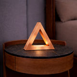 Copper finish triangular table lamp by WOOM, casting a warm glow on a dark marble-top wooden bedside table.