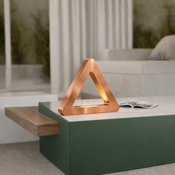 Copper finish triangular LED lamp by WOOM, casting a warm glow on a marble coffee table in a modern living room.