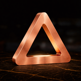 Copper finish triangular LED lamp by WOOM, with a brushed texture, sitting on a dark marbled surface.