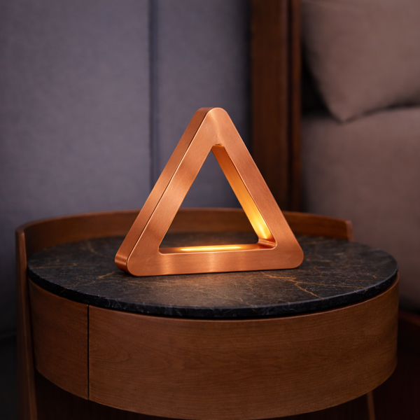 Copper triangular LED lamp by WOOM, glowing softly on a round oak table with a black marble top.
