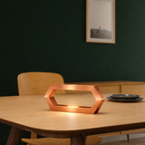 Copper finish hexagon LED lamp by WOOM, glowing warmly on a light wooden table with dark plates.