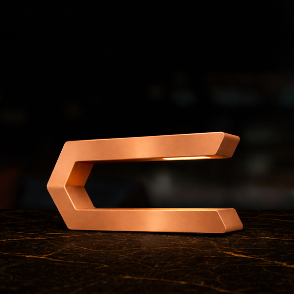 Copper finish C-shaped LED lamp by WOOM with light glowing from under the top bar, on a dark marble surface.