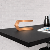 Copper finish C-shaped LED lamp by WOOM emitting a warm glow on a black desk against a white brick wall.