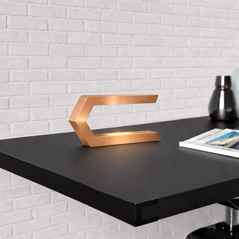 Copper finish C-shaped LED lamp by WOOM emitting a warm glow on a black desk against a white brick wall.