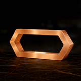 Copper finish hexagon LED lamp by WOOM with a glowing white light, sitting on a dark marbled surface.