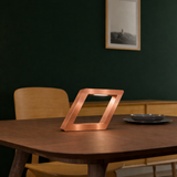 Copper finish rotatable square lamp by WOOM, with a geometric design and internal light on a dark wooden table.