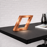 Copper finish handcrafted oak rotatable square wooden lamp by WOOM, illuminating a dark desk against a white brick wall.