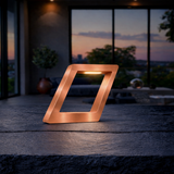 Copper finish geometric wooden table lamp by WOOM, with a warm internal glow, on a dark textured surface.
