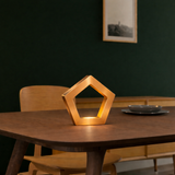 Copper finish pentagon LED lamp by WOOM, glowing warmly on a wooden dining table.
