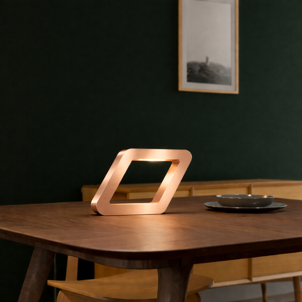 Copper finish rotatable square wooden table lamp by WOOM, emitting a warm glow on a dark wooden table against a deep green wall.