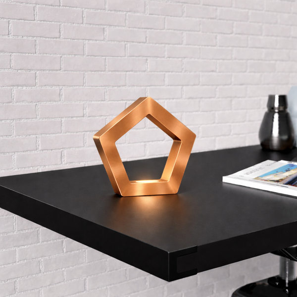Copper finish pentagon wooden LED lamp by WOOM, glowing from within on a dark shelf against a white brick wall.