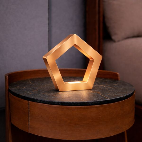 A copper finish pentagon LED lamp by WOOM, glowing on a dark marble-top wooden side table.