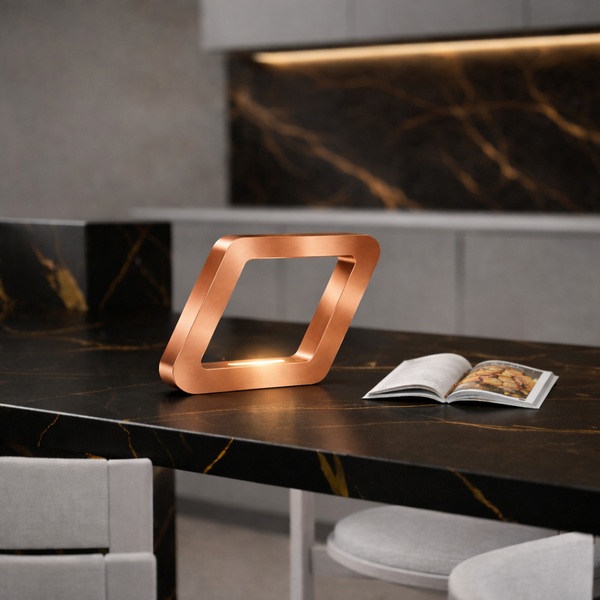 Copper finish rotatable square wooden lamp by WOOM, emitting a warm glow on a black marble countertop, beside an open magazine.