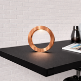 Copper finish handcrafted wooden oak rotatable circle table lamp by WOOM, glowing on a black table against a white brick wall.