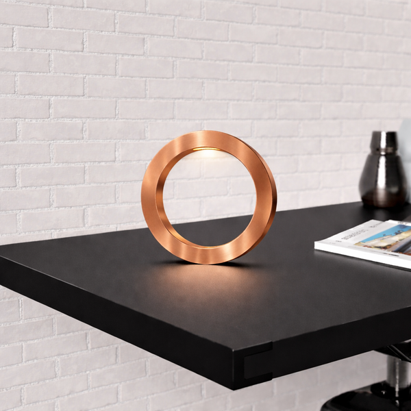 Copper finish handcrafted wooden oak rotatable circle table lamp by WOOM, glowing on a black table against a white brick wall.