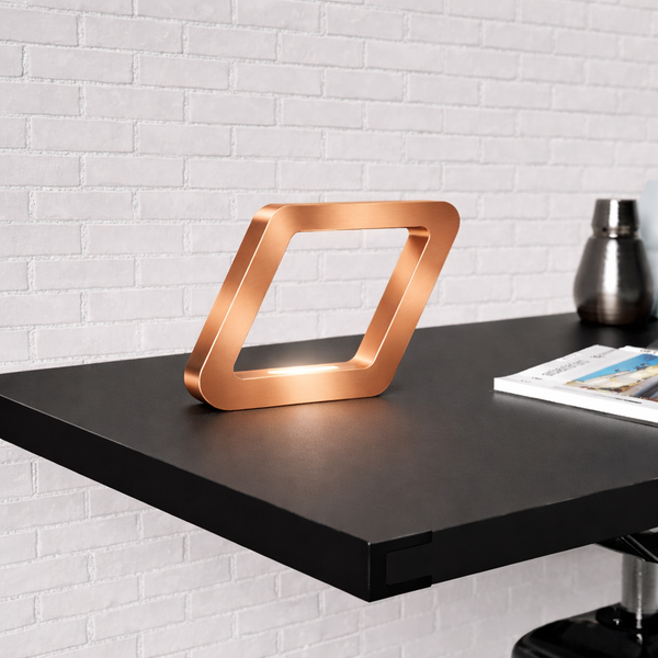 Copper-finish square-shaped table lamp by WOOM, illuminated on a dark desk against a white brick wall.