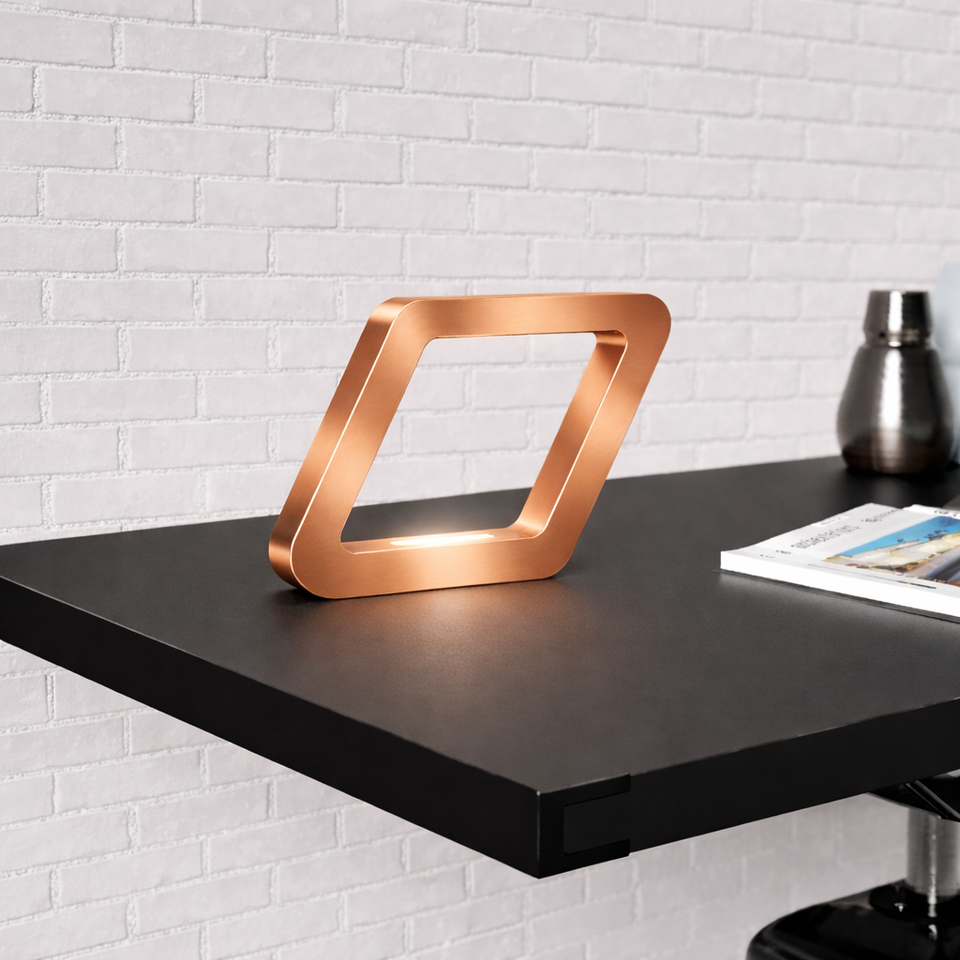 Copper-finish square-shaped table lamp by WOOM, illuminated on a dark desk against a white brick wall.