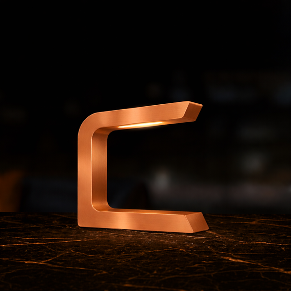 Copper finish C-shaped table lamp by WOOM, emitting a warm glow, set on a dark marble surface.