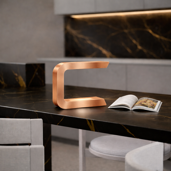 Copper finish C-shaped lamp by WOOM, glowing from within, on a dark marble counter next to an open book.