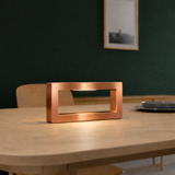 Copper finish rectangular wooden LED lamp by WOOM, emitting a warm light on a light wooden table.
