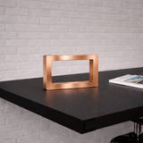 A rectangular copper-finish wooden LED lamp by WOOM, resting on a dark table against a white brick wall, with a magazine nearby.