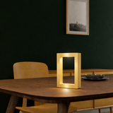 Gold finish rectangular wooden table lamp by WOOM with an illuminated hollow center on a dark dining table.