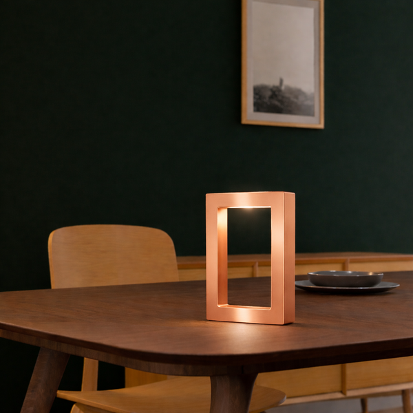 Copper finish rectangular table lamp by WOOM with an illuminated opening, on a dark wooden table.