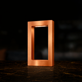 Copper finish rectangular lamp by WOOM with an illuminated interior, on a dark marble surface with gold veins.