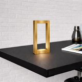 Gold finish rectangular table lamp by WOOM glowing on a black shelf against a white brick wall.