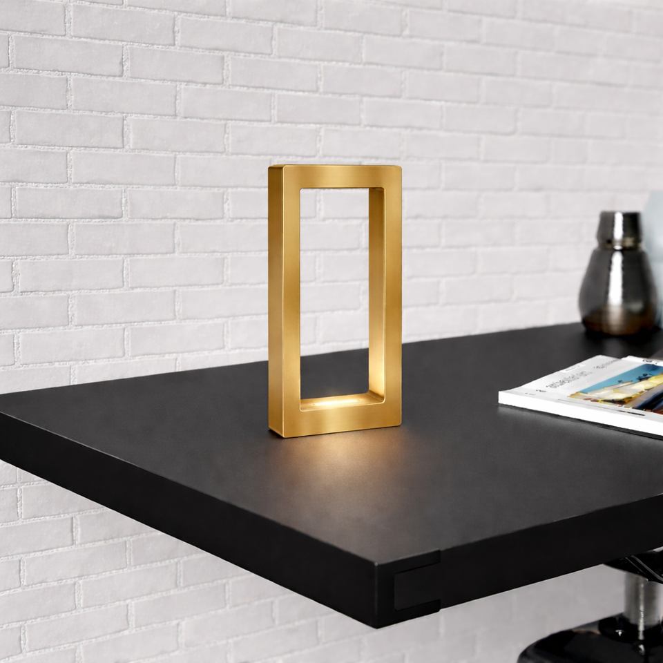 Gold finish rectangular table lamp by WOOM glowing on a black shelf against a white brick wall.
