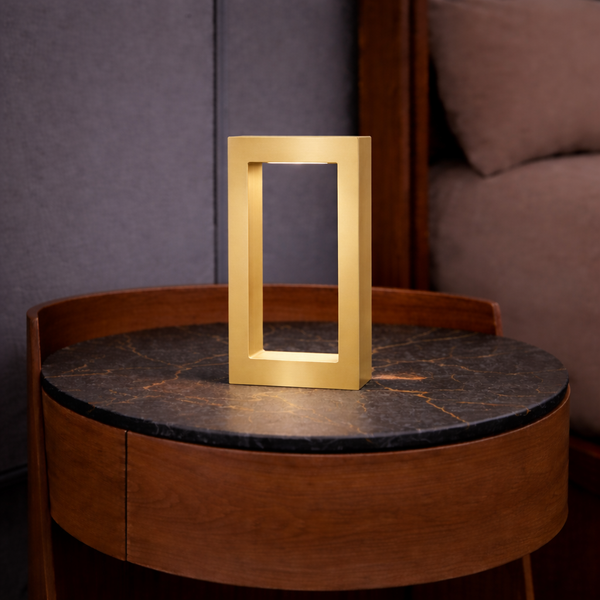 Gold finish oak rectangular table lamp by WOOM, with an open center and internal light, on a marble-top bedside table.