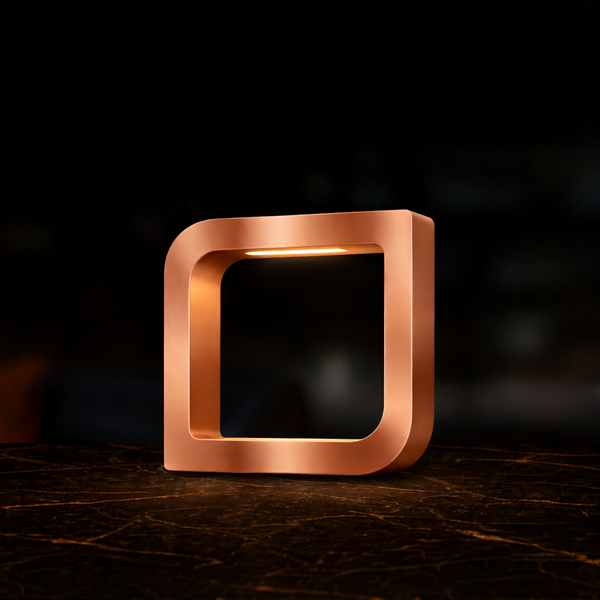 Copper finish square LED lamp by WOOM with rounded corners and an internal light, on a dark marble table.