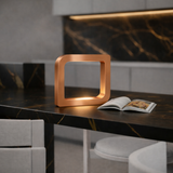 Rounded square copper finish LED table lamp by WOOM, emitting a warm glow on a dark marble countertop with gold veins, beside an open magazine.