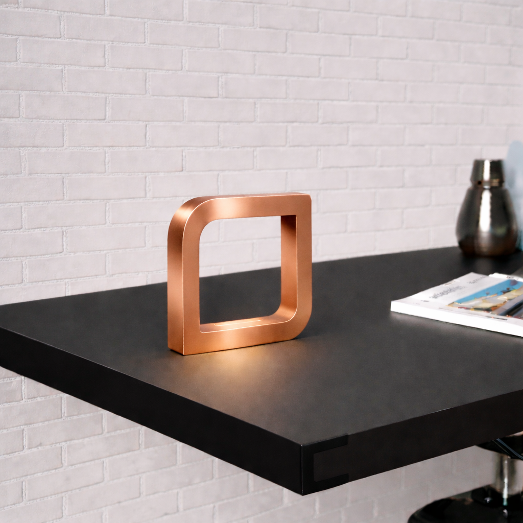 Copper finish square LED lamp by WOOM, with rounded corners, on a dark table in front of a white brick wall.