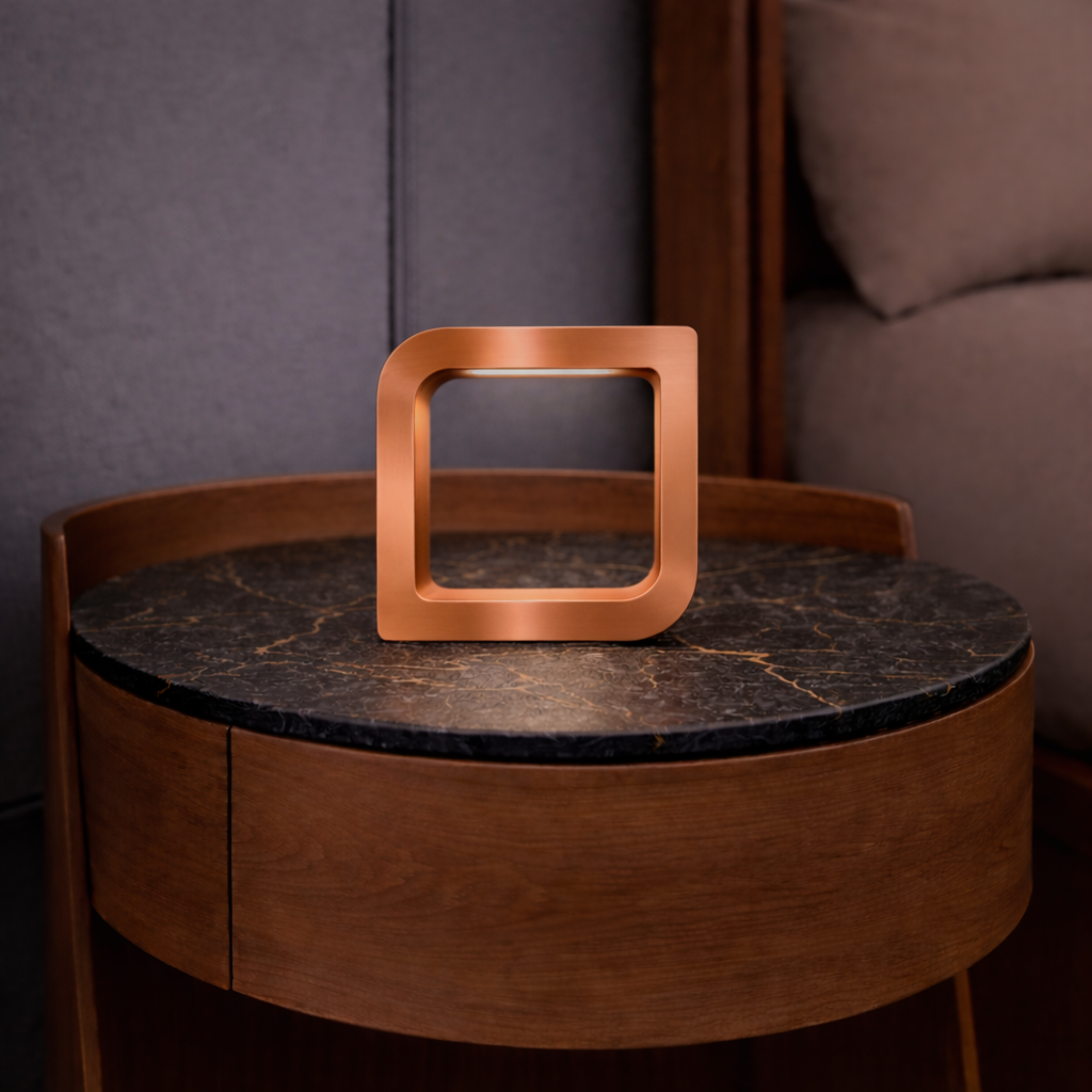 Square copper finish LED lamp by WOOM glowing softly on a dark marbled and wooden side table.