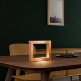 Copper finish square lamp by WOOM, softly glowing on a dark wooden table.