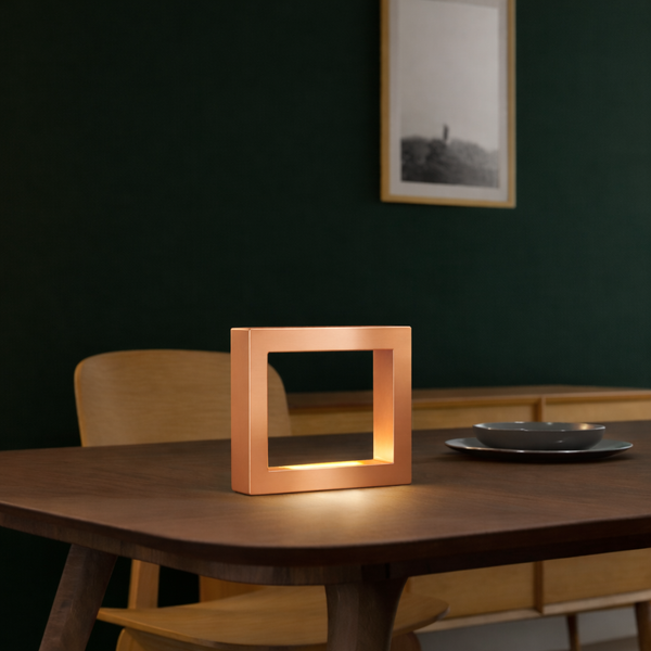 Copper finish square lamp by WOOM, softly glowing on a dark wooden table.