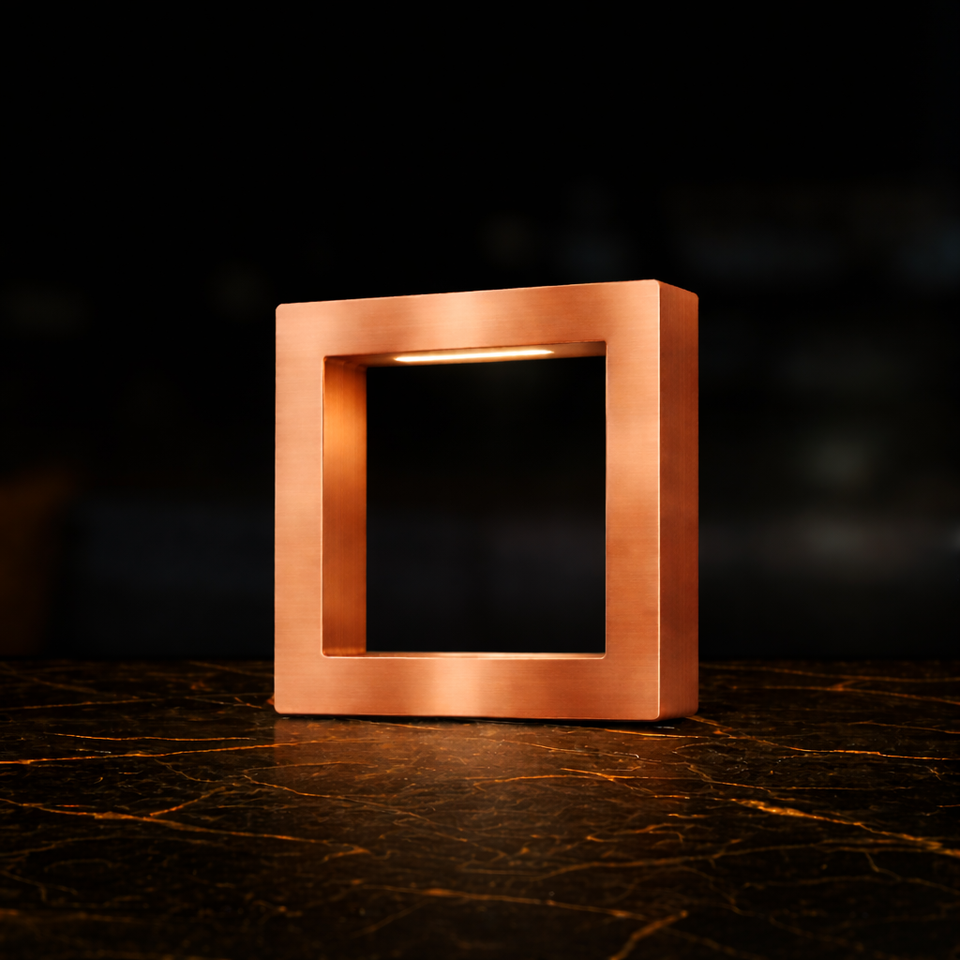 Copper finish square table lamp by WOOM, hollow design featuring a warm LED light, on a dark marble table with golden veins.