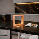 Copper Finish square wooden lamp by WOOM on a dark marble counter with golden veins next to an open book.
