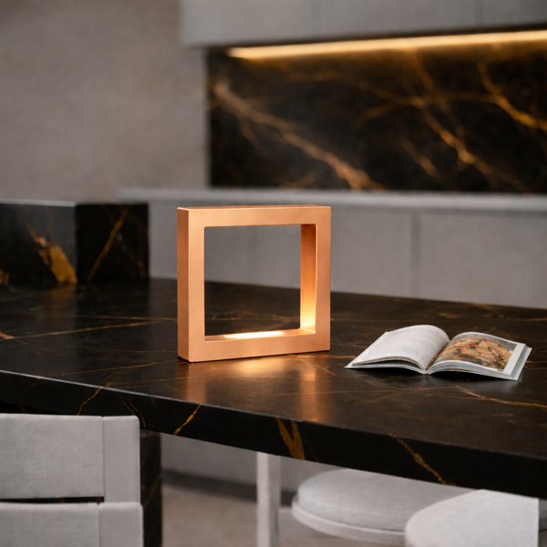 Copper Finish square wooden lamp by WOOM on a dark marble counter with golden veins next to an open book.