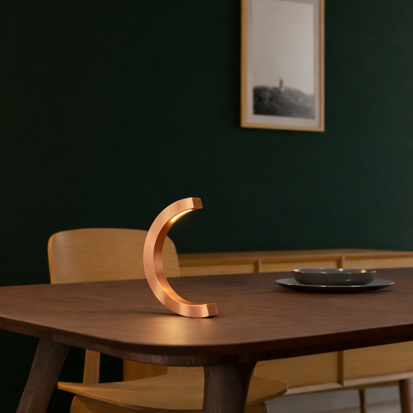 Copper finish oak C-curved desk lamp by WOOM glowing warmly on a wooden dining table.