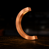 Copper finish C-curved wooden desk lamp by WOOM with warm inner lighting, on a dark textured table.