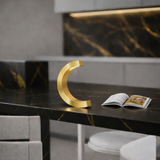 Gold finish C-curved wooden desk lamp by WOOM, glowing on a dark countertop with a magazine.