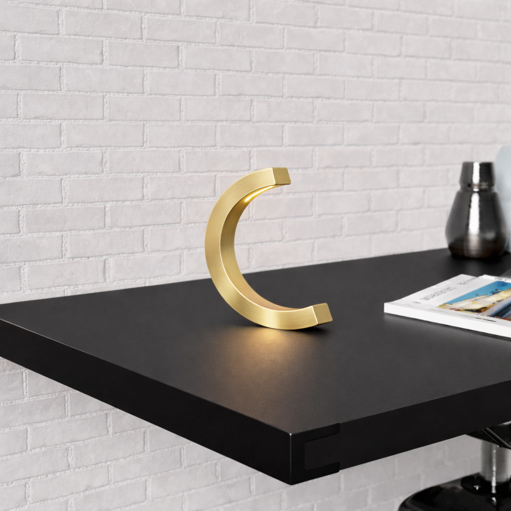 Gold finish C-curved wooden desk lamp by WOOM, glowing softly on a black shelf with a white brick wall.