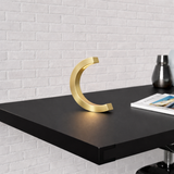 Gold finish C-curved wooden desk lamp by WOOM, glowing softly on a black shelf with a white brick wall.