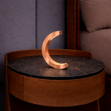 Copper finish handcrafted oak C-curved desk lamp by WOOM with integrated light, on a dark marble side table.