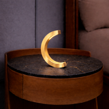 Gold finish C-curved oak desk lamp by WOOM on a dark marble-top side table.