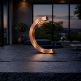 Copper finish C-curved wooden desk lamp by WOOM, with an internal warm glow on a dark textured surface.