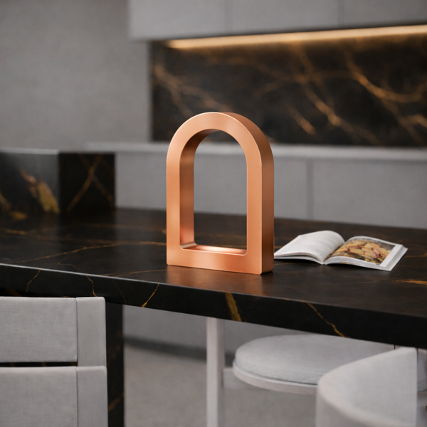 Copper finish U-shape curved desk lamp by WOOM, displayed on a dark marble kitchen island with an open book.